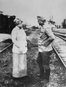 Bly is on the left, wearing a light suit with a long skirt. On the right is an Austrian officer. They are standing near railway tracks, and both have their hands in their pockets.