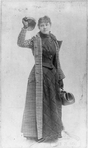 Bly is wearing a travelling outfit of a dress with a long skirt and tight bodice. Over it is a floor length check coat. She has a bag in one hand a is raising her cap with the other.