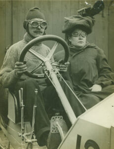 Two white people sat in an early racing car. Then man driving it is wearing a sweater, a balaclava and googles. The woman next to him is in a large overcoat, goggles and what looks like a very impractical hat.