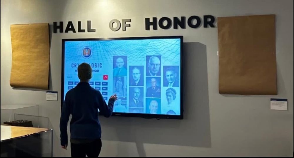 Photo of the Hall of Honor at the National Cryptologic Museum in Maryland. Someone is standing at an interactive display. On either side of it, large sheets of plain brown paper are hung up, covering two display panels.